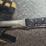 A worn duct board knife with the serrated edge sharpened off, lying on galvanized next to a pair of work gloves